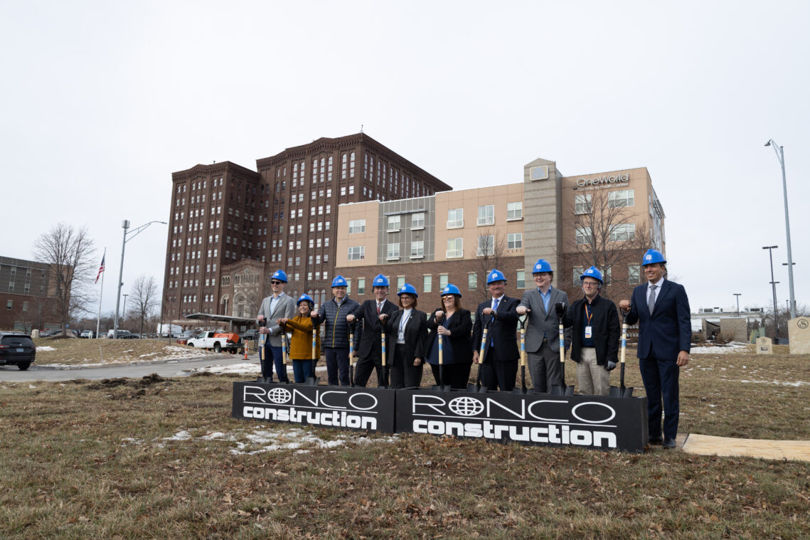 A line of people wearing hard hats hold shovels as part of a groundbreaking ceremony. In the background are existing buildings on the main OneWorld campus.